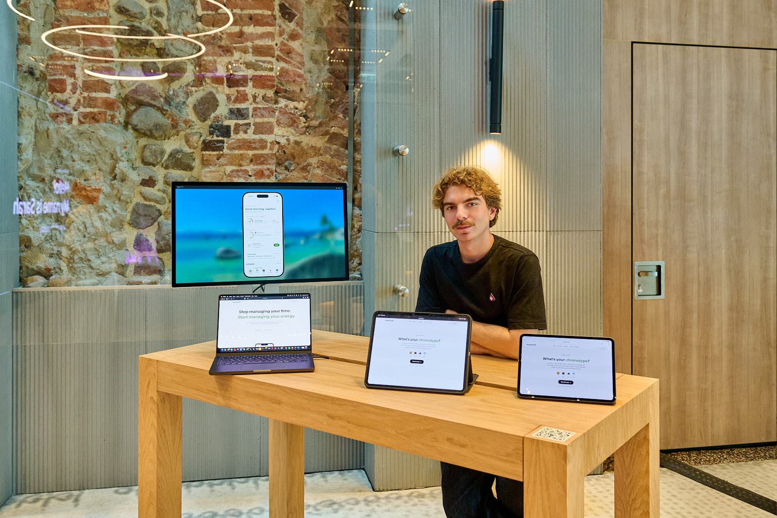 Joachim z AcademiX przy stanowisku: na stole MacBook i dwa iPady z quizem chronotypu, na monitorze z tyłu ekran aplikacji